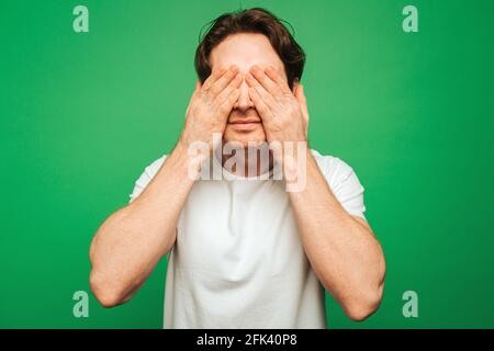 Young man covers eyes up with hands, isolated on green background Stock Photo