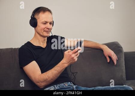 The man in headphones and with a smartphone sits on the couch. Stock Photo