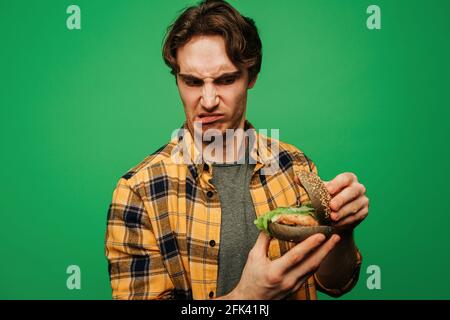 Young man is disappointed because of spoiled burger, isolated on green background Stock Photo