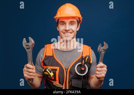 Smiling builder in a helmet with wrenches, isolated over blue background Stock Photo