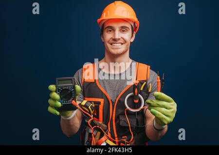 Happy builder is holding digital multimetert in gloves, isolated over blue background Stock Photo
