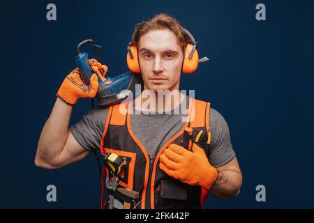 Young builder is serious while having equipment in arm, isolated over blue background Stock Photo