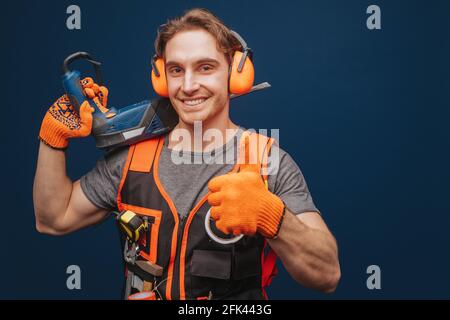 Young builder is showing thumb up while having equipment in arm, isolated over blue background Stock Photo