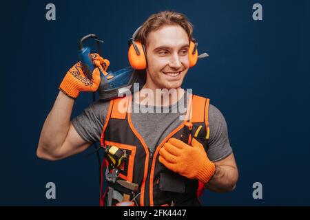 Young builder is smiling while having equipment in arm, isolated over blue background Stock Photo