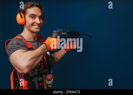 Young builder is drilling the wall and smiling, isolated over blue background Stock Photo