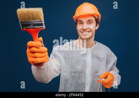 Young builder is holding brush to paint wall and smiling, isolated over blue background Stock Photo