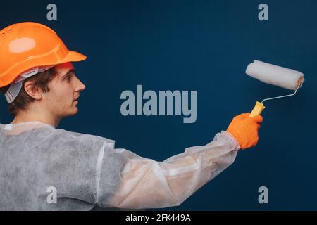 Builder is using roller to paint wall, isolated over blue background Stock Photo