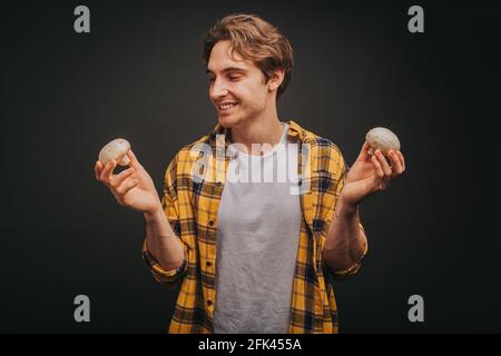 Young blond man in yellow shirt is holding mushrooms in both hands, isolated over black background Stock Photo