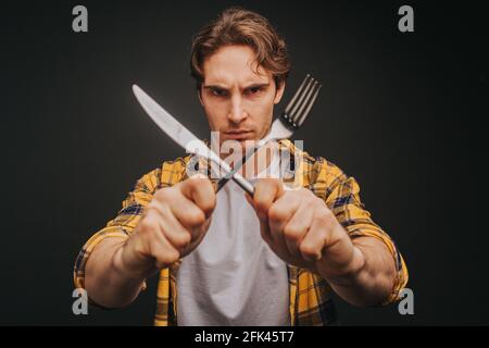 Young blond man in yellow shirt is smiling while holding fork and knife making stop, isolated over black background Stock Photo
