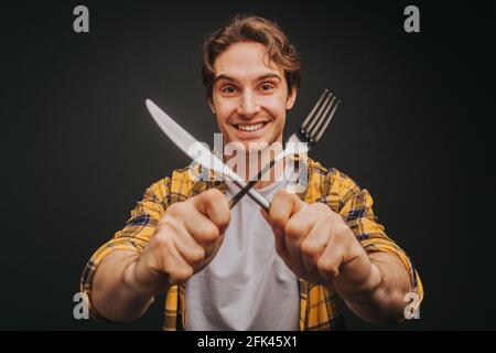 Young blond man in yellow shirt is smiling while holding fork and knife, isolated over black background Stock Photo
