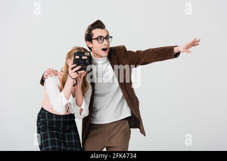 amazed man pointing with hand near girlfriend taking photo on vintage camera isolated on grey Stock Photo