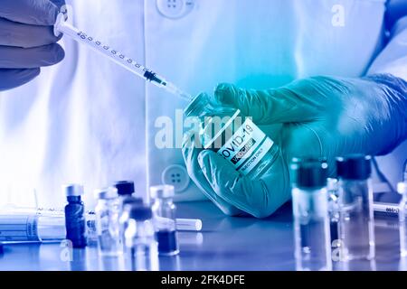 a healthcare or laboratory worker, wearing surgical gloves, fills syringe from a vial of simulated covid-19 vaccine, on a blue table full of laborator Stock Photo