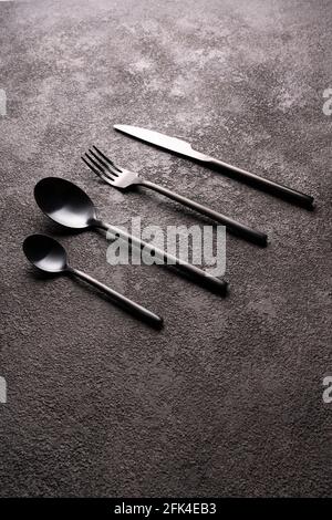 Black cutlery-fork, spoon, knife, on a dark background in the center. Minimalistic still life, vertical photo. Stock Photo