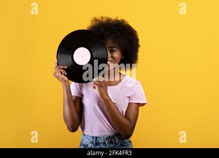 Music lover. Happy african american woman holding vinyl record and covering eye, posing over yellow studio background Stock Photo