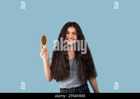 Portrsit of beautiful Indian teen girl with long dark hair holding wooden brush and smiling at camera on blue background Stock Photo
