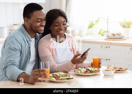 Millennial Black Man And Woman Having Breakfast And Using Smartphone In Kitchen Stock Photo