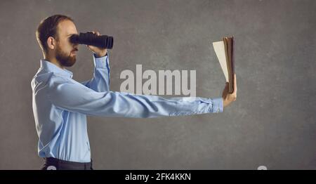 Side view of young guy with binoculars reading book he is holding in long extended arm Stock Photo