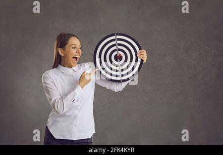 Happy young businesswoman holding dartboard target with red dart right in bullseye Stock Photo