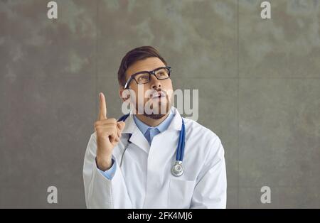 Young doctor in glasses and lab coat with stethoscope thinking with one finger pointing up Stock Photo