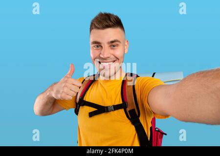 Happy male traveler with backpack showing thumb up gesture while making selfie over blue studio background