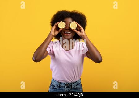 Natural vitamins for skin detox. Positive black woman with lemon halves in front of her eyes posing on yellow background Stock Photo