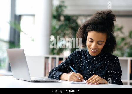 Focused young african american woman with headset, dressed in formal shirt, manager or freelancer, sitting at table in office, working at laptop, taking notes, smiling Stock Photo