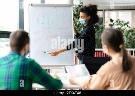 Multiracial employees gathered together at office, successful smart young african american business lady wearing medical mask, conducts brainstorm with colleagues, explains business strategy and plan Stock Photo