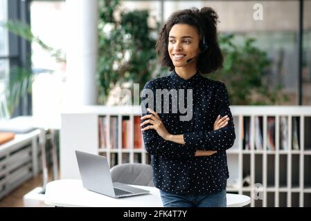 Portrait of a pretty, pleasant young african american woman, with headset and in stylish wear, freelance, call center worker or support operator, stands near workplace, looks away, arms crossed, smile Stock Photo