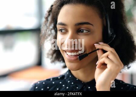 Close-up headshot of pleasant charming young african american curly haired woman in headset and stylish shirt, female call center worker or support operator, looking to the side, smiling friendly Stock Photo