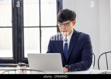 Side view handsome young businessman in eyewear working with computer remotely, sitting at wooden table in kitchen. Pleasant happy man communicating i Stock Photo