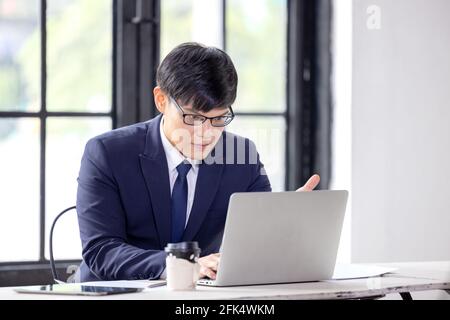 Side view handsome young businessman in eyewear working with computer remotely, sitting at wooden table in kitchen. Pleasant happy man communicating i Stock Photo