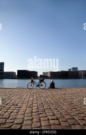 Blonde woman sitting on boardwalk and looking a the water in harbor with a bicycle standing next to her. Seen from behind and sun reflecting in water Stock Photo