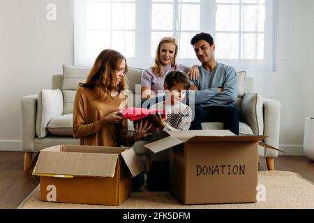 Cute little caucasian boy putting things like clothes in cardboard box for donation with help of teenage sister while happy mother and father watching them at home Stock Photo