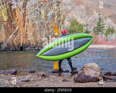 male kayaker is carrying a whitewater inflatable kayak on a shore of mountain river in early spring - Poudre River in northern Colorado Stock Photo