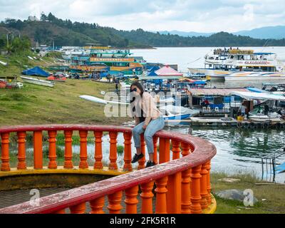 Guatapé, Antioquia, Colombia - April 4 2021: Latina Woman Posing on an Orange Balcony Against a Backdrop of a Lake Coast full of Boats and Ships full Stock Photo