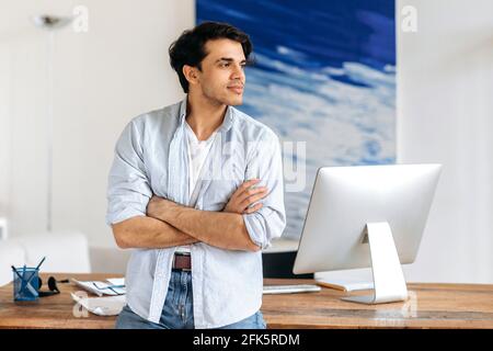 Successful attractive young mixed race male, stylishly dressed designer, freelancer or manager, standing near his desk with crossed arms and looking away while thinking about business idea or vacation Stock Photo
