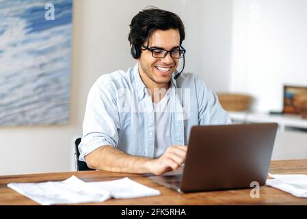 Happy successful confident young hispanic man in headphones, call center employee or freelancer, works remotely on laptop, communicating via video communication with employees, browses internet, smile Stock Photo