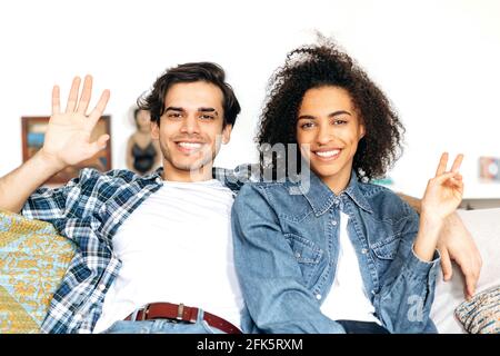 Portrait of interracial friendly happy young family couple sitting on sofa, stylishly dressed, looking waving hands at the camera, smiling, spend time together. Relationship between multiethnic people