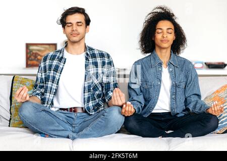 Relaxed multiracial married couple in love, sitting in lotus position on the couch at home wearing casual wear, relieve tension relaxing with eyes closed, meditate, guy and girl spend time together Stock Photo