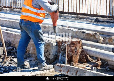 A road worker in an orange reflective vest works with a jackhammer to smash an old reinforced concrete structure around a rusty support. Copy space. Stock Photo