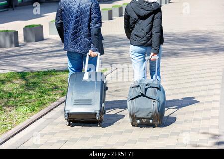 Two people, a man and a woman, walk along the cobbled sidewalk, carrying a travel bag and a suitcase on wheels behind them. Stock Photo