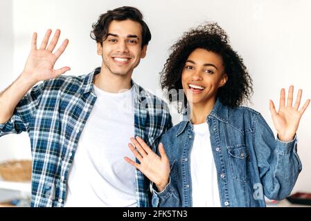 Joyful guy and girl posing at camera. Portrait of friendly joyful young multiracial couple standing in casual stylish clothes, are greet with hand gesture at webcam, look at camera, smiling happily Stock Photo