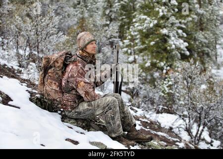 gun hunter wearing camouflage and sitting in snowy scene holding rifle Stock Photo