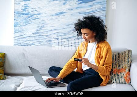 Online shopping concept. Young beautiful african american woman in casual stylish clothes sits on the couch, makes purchases online by laptop, fills in credit card details to pay for goods or delivery Stock Photo