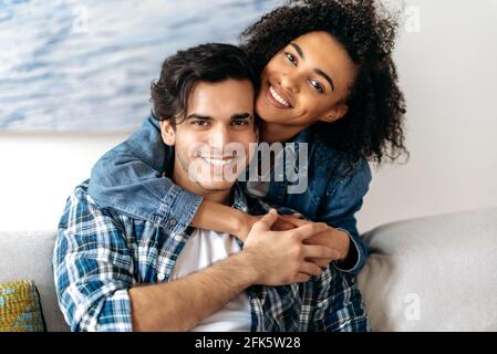 Portrait joyful happy mixed race couple, pretty african american curly girl tenderly hugging her hispanic boyfriend sitting on sofa, wearing casual clothes in living room, looking at camera, smiling Stock Photo