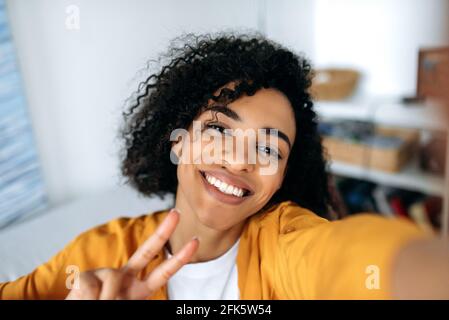 Cute beautiful joyful curly-haired African American girl, teenager, posing for selfie on smartphone, fooling around, having fun, looking at phone camera, smiling happily Stock Photo