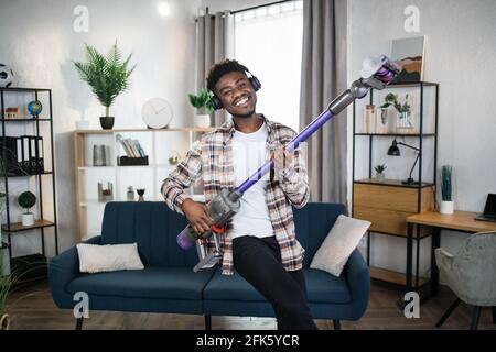 Cheerful afro american man imitating play on guitar using handheld vacuum cleaner. Happy young man listening music in headphones and cleaning house with modern device. Stock Photo