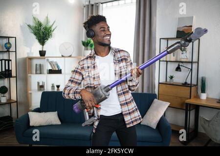 Cheerful afro american man imitating play on guitar using handheld vacuum cleaner. Happy young man listening music in headphones and cleaning house with modern device. Stock Photo