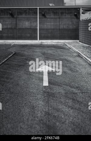 Black and white shot of an asphalt road with a selective focus on a white contrast arrow on it as a road marking leading to a concrete wall and a turn Stock Photo