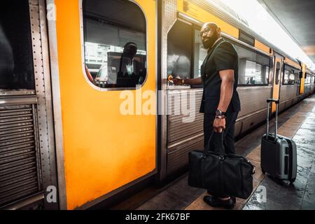 A wide-angle view of a mature bald bearded black man entrepreneur in a suit and with luggage bags, pushing the button to open a vivid yellow door of a Stock Photo